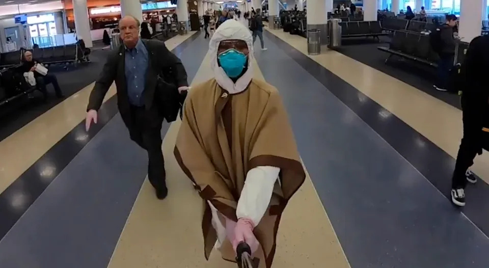 A person in a brown cloak with a hood and face mask walks through an airport, holding a selfie stick