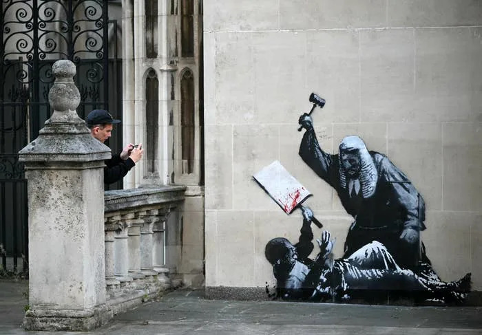 A person photographs a Banksy street art piece of a judge hitting a figure with a gavel, symbolizing justice and authority