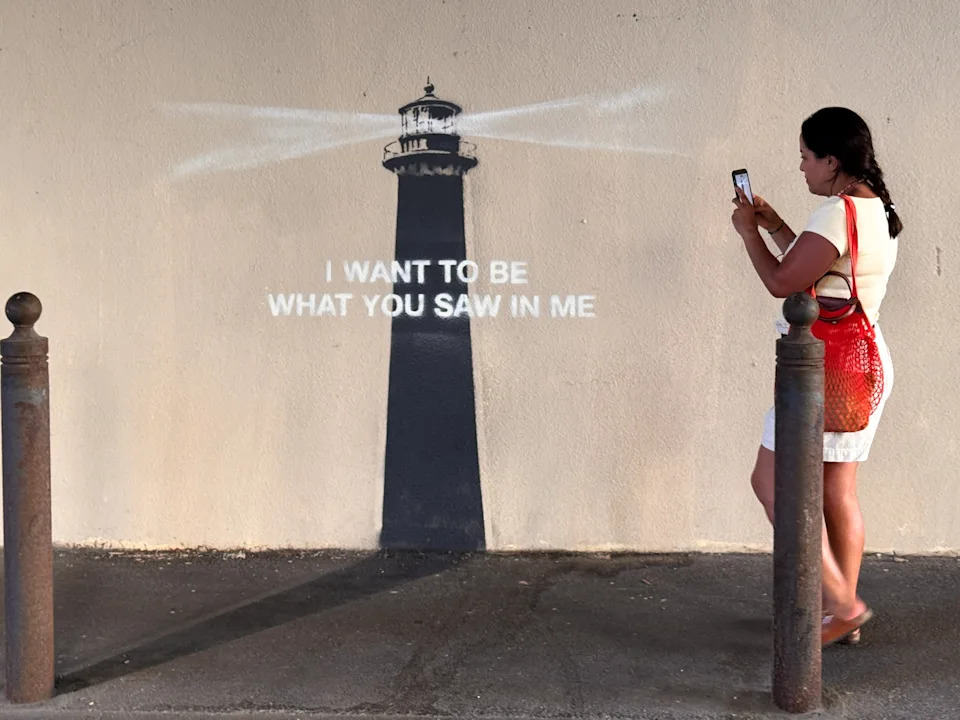 A woman takes a photo of a stencil art piece on a wall featuring a lighthouse and the text "I WANT TO BE WHAT YOU SAW IN ME."