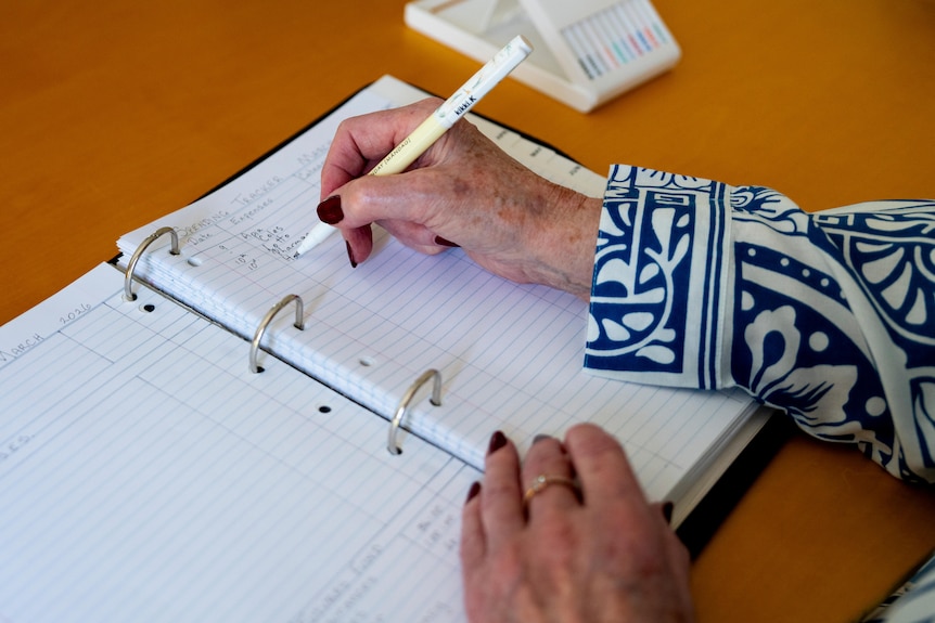 Suzenne's woman's hand writing with a white pen on a lined piece of paper in a binder on a brown table.