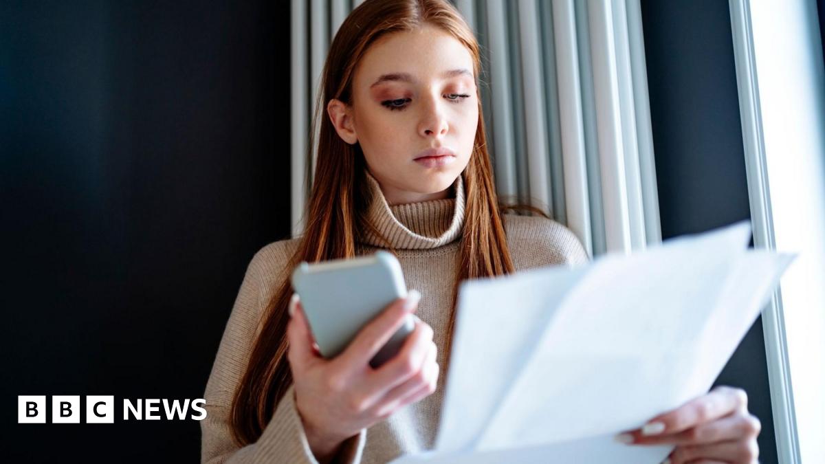 Young woman with red hair, wearing beige jumper holds smartphone in one hand and household bills in the other