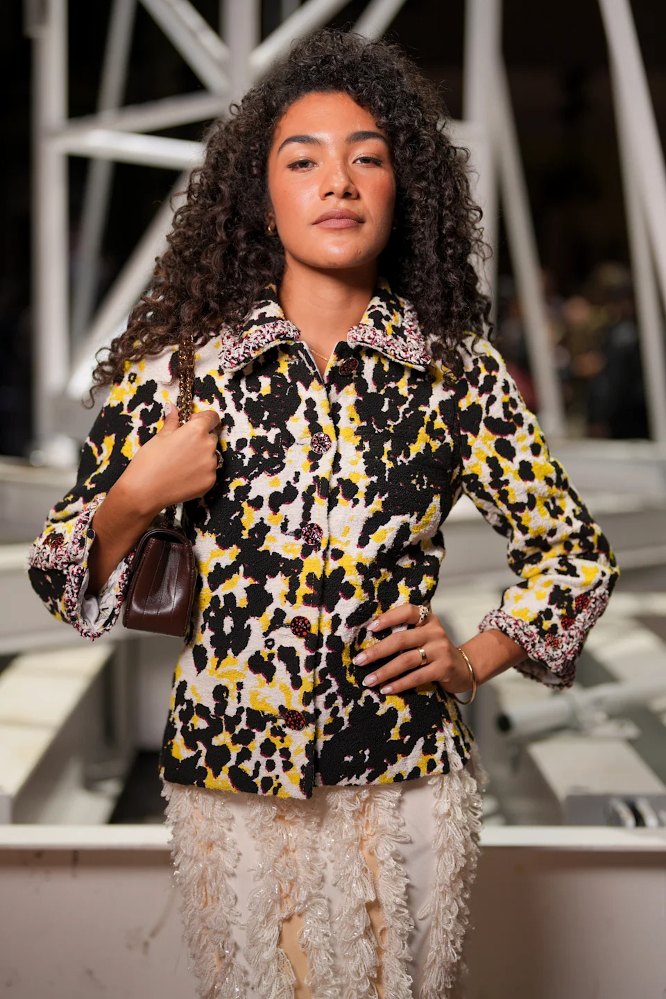 Person in patterned jacket and textured skirt on a red carpet, carrying a small handbag, with curled hair, posing confidently