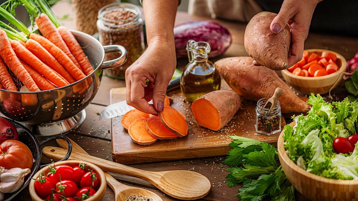 Hands slicing sweet potatoes on a wooden cutting board surrounded by fresh carrots, lettuce, tomatoes, and olive oil in a rustic kitchen.