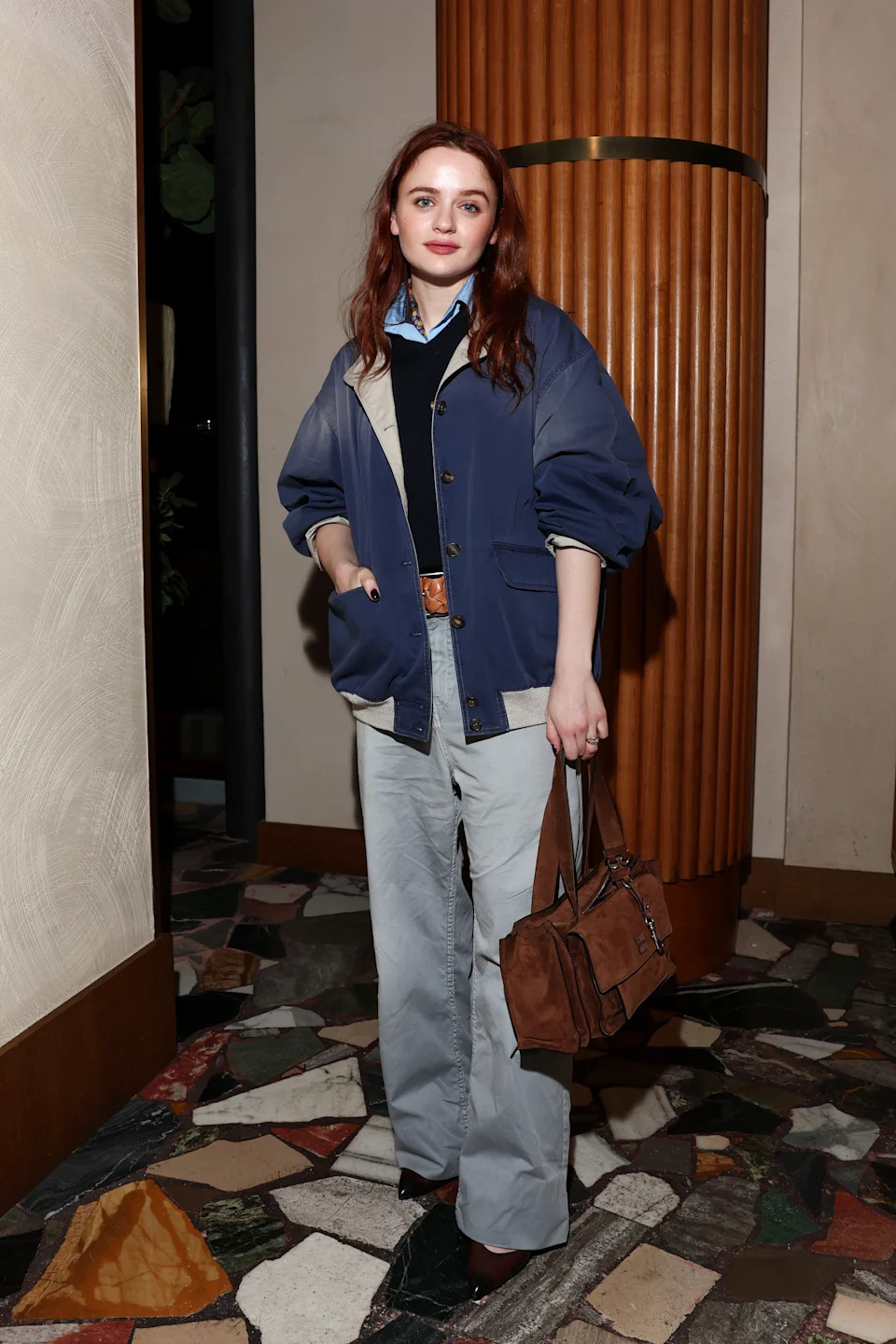 A person in stylish casual wear with a blue jacket and gray pants poses confidently, holding a brown bag, at a formal event indoors