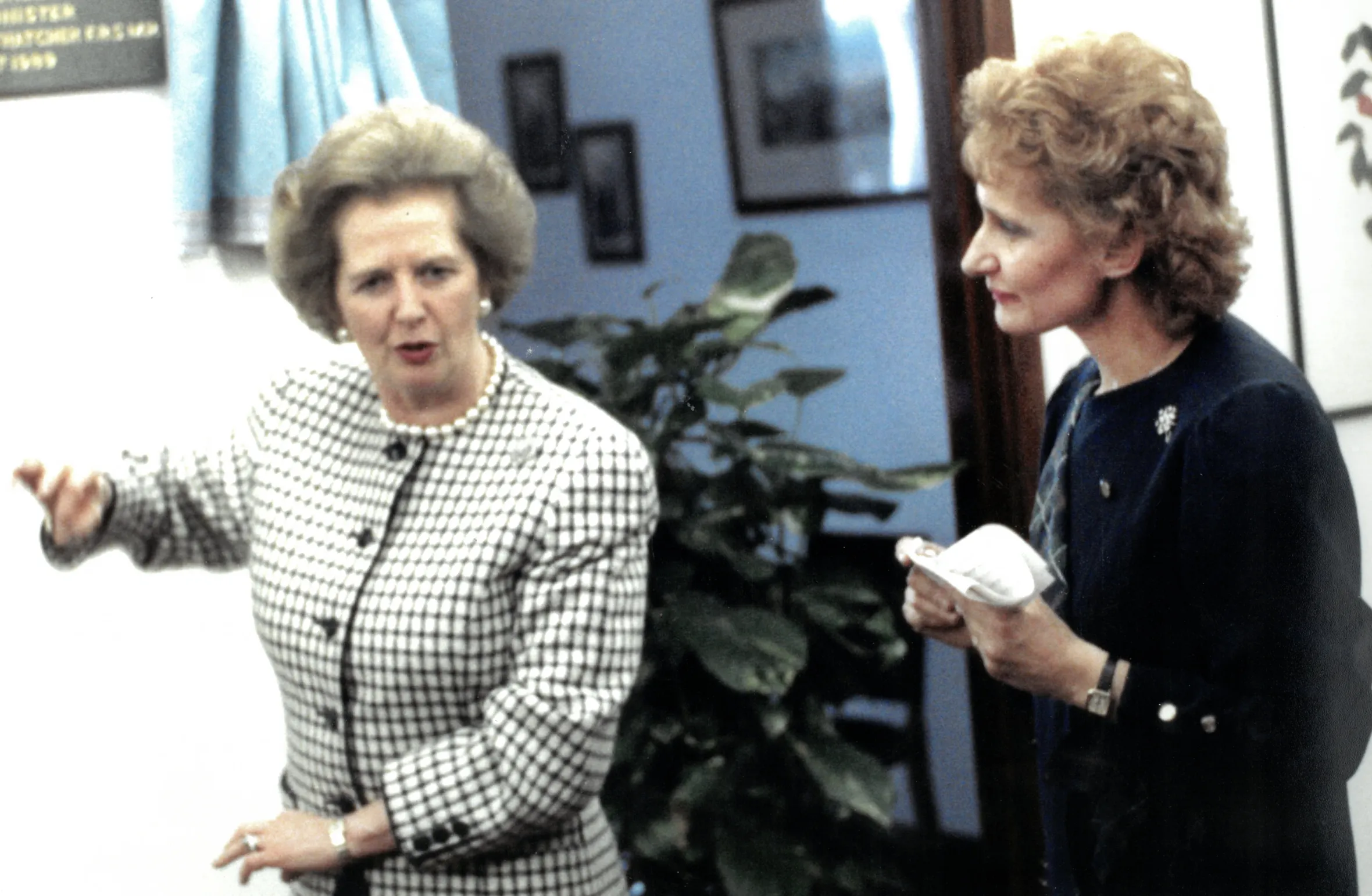 Margaret Thatcher in a checkered jacket gestures while speaking to June Powell.