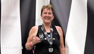 Carole Munro smiles at the camera wearing a medal and holding a patch. She is standing in front of a black and white striped backdrop and is wearing a black vest top.