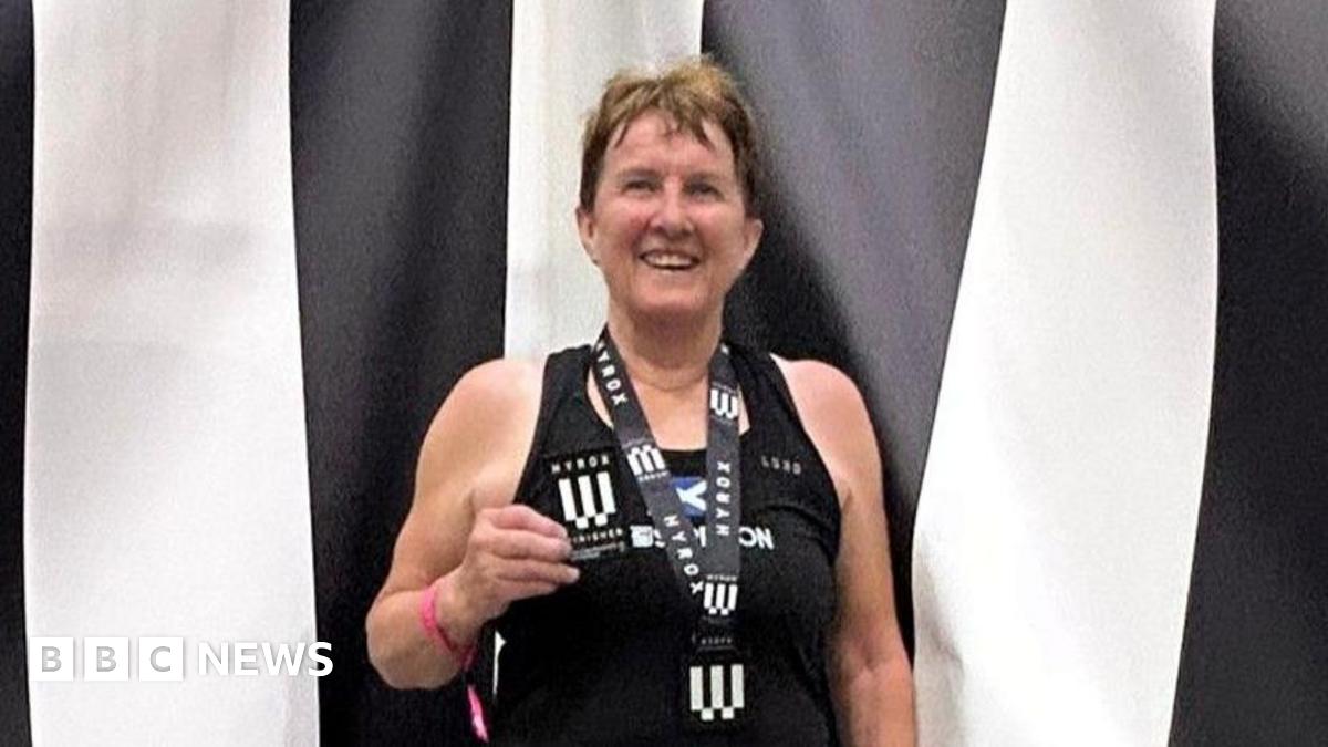 Carole Munro smiles at the camera wearing a medal and holding a patch. She is standing in front of a black and white striped backdrop and is wearing a black vest top.