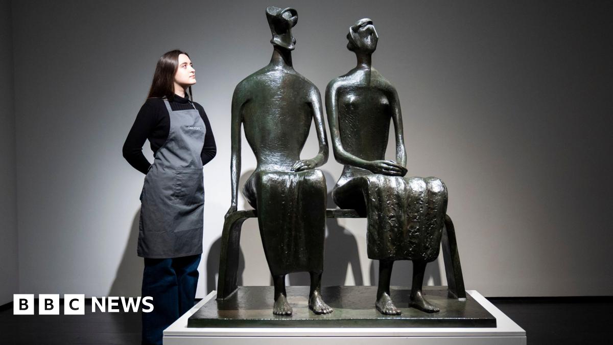 A bronze sculpture of a couple sitting on a bench. A woman in a black top and grey apron stands next to the sculpture with her hands behind her back, looking at it.