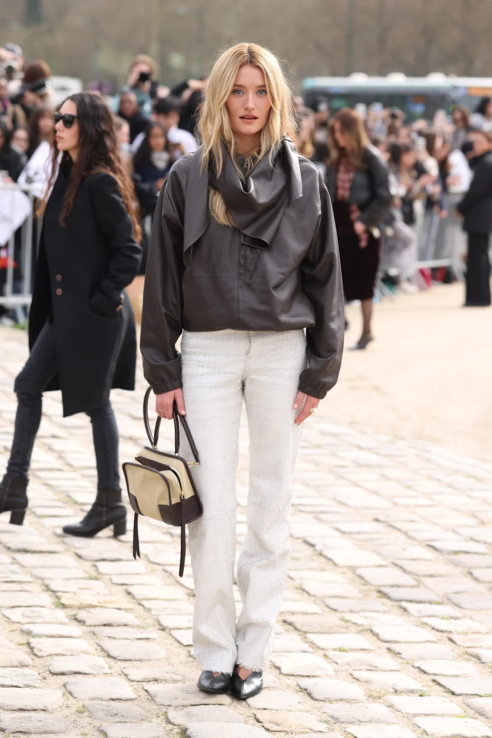 Person standing on a cobblestone path wearing a leather jacket, white pants, and holding a two-tone bag. Crowd in the background