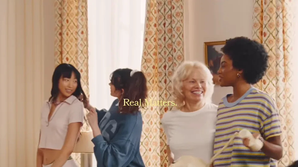 Four women laughing and talking indoors, with mid-action hair styling. Text reads: "Real Matters."