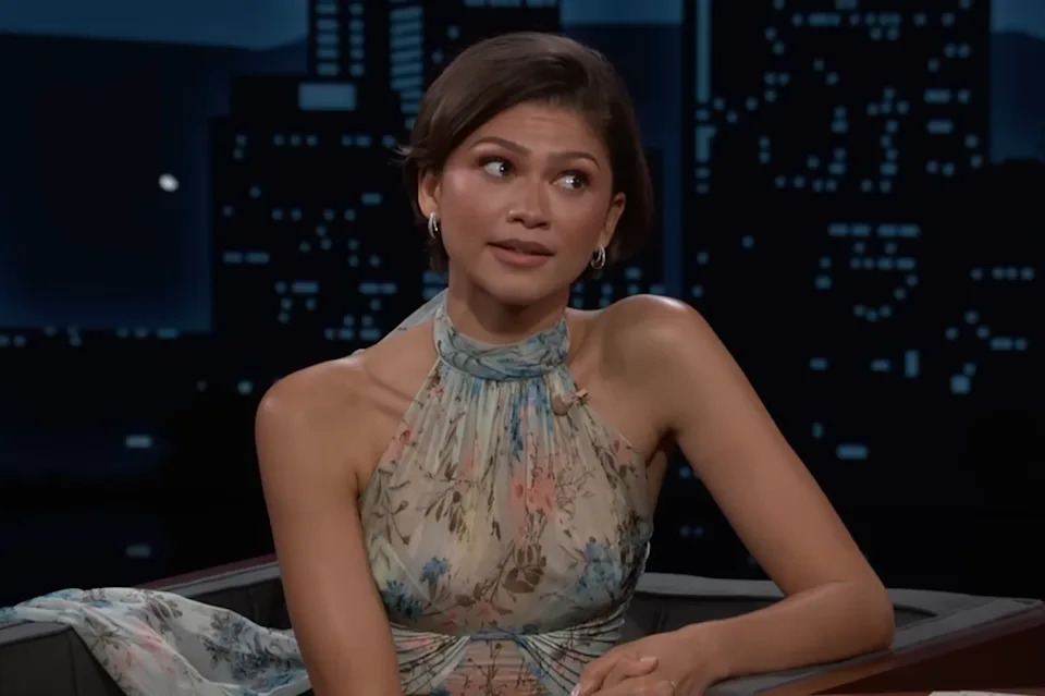 A celebrity sits on a talk show set, wearing a floral halter dress with a soft pattern, gesturing slightly with a thoughtful expression