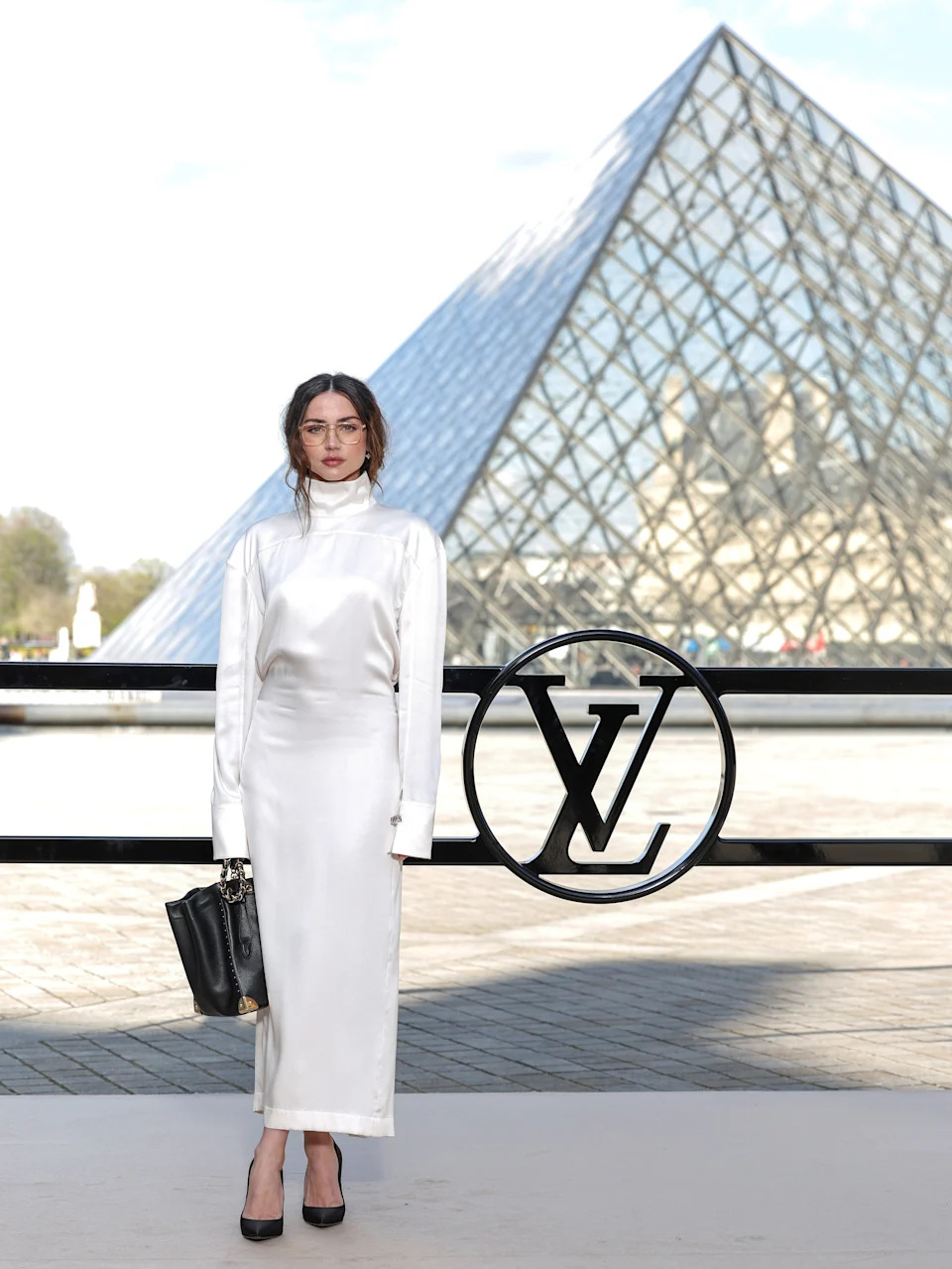 Person in a sleek long-sleeve dress stands in front of a large glass pyramid, holding a bag, likely at a fashion event