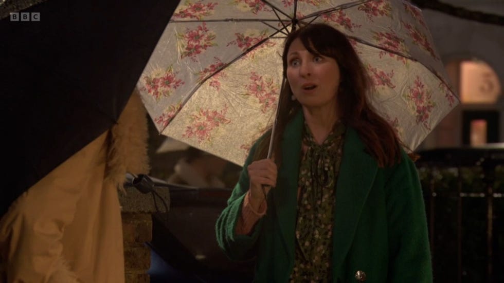 eastenders honey mitchell outdoors holding a floral umbrella and wearing a green coat