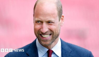 Prince William, wearing a navy suit and red tie, smiles