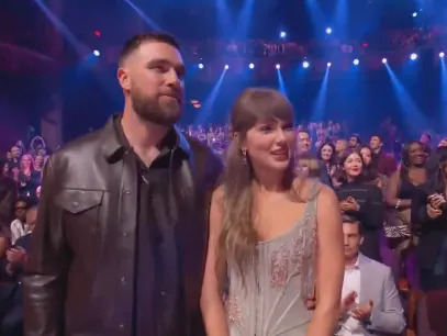 A man in a leather jacket and a woman in an elegant dress are standing together at an event, with an audience in the background