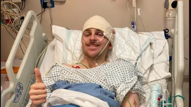 A man in a hospital bed smiling and giving a thumbs up. His head is bandaged and he wears a hospital gown.
