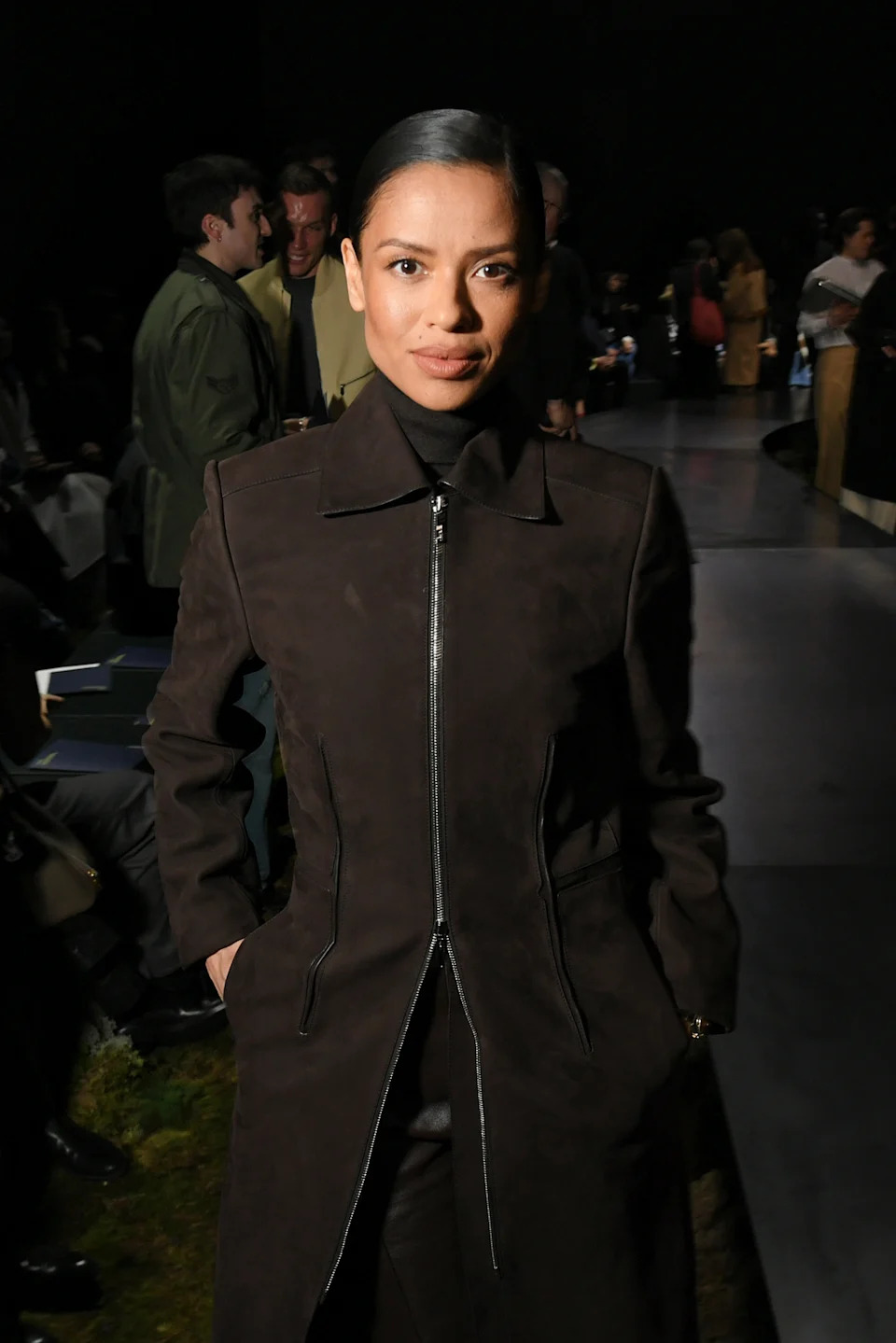 A person in a sleek, dark coat poses confidently at a fashion event, surrounded by attendees