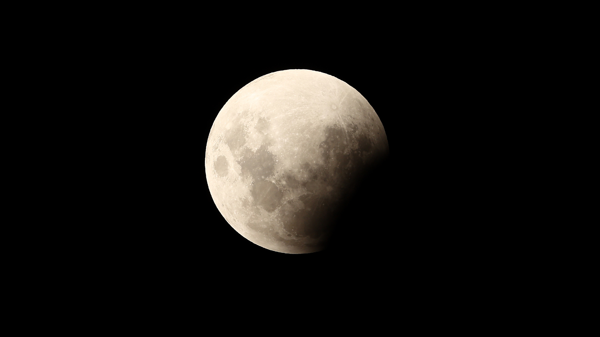 A full moon is photographed in a black sky with its lower-right edge obscured by Earth's shadow during an eclipse. Dark patches called lunar maria mark its surface, where lava once flooded ancient impact craters.