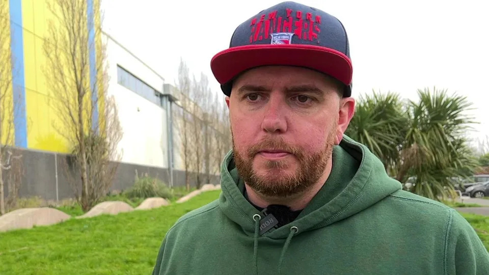 David Haddock stood outside the Plymouth Life Centre. He is wearing a green hoody and black and red hat. He has a stern look on his face as he looks slight away from the camera.