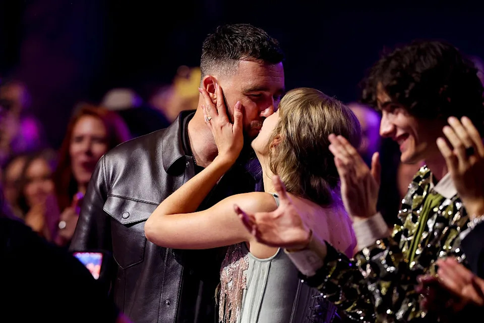 A couple shares a kiss at an event, surrounded by clapping attendees. They are in stylish formalwear, and the atmosphere is celebratory