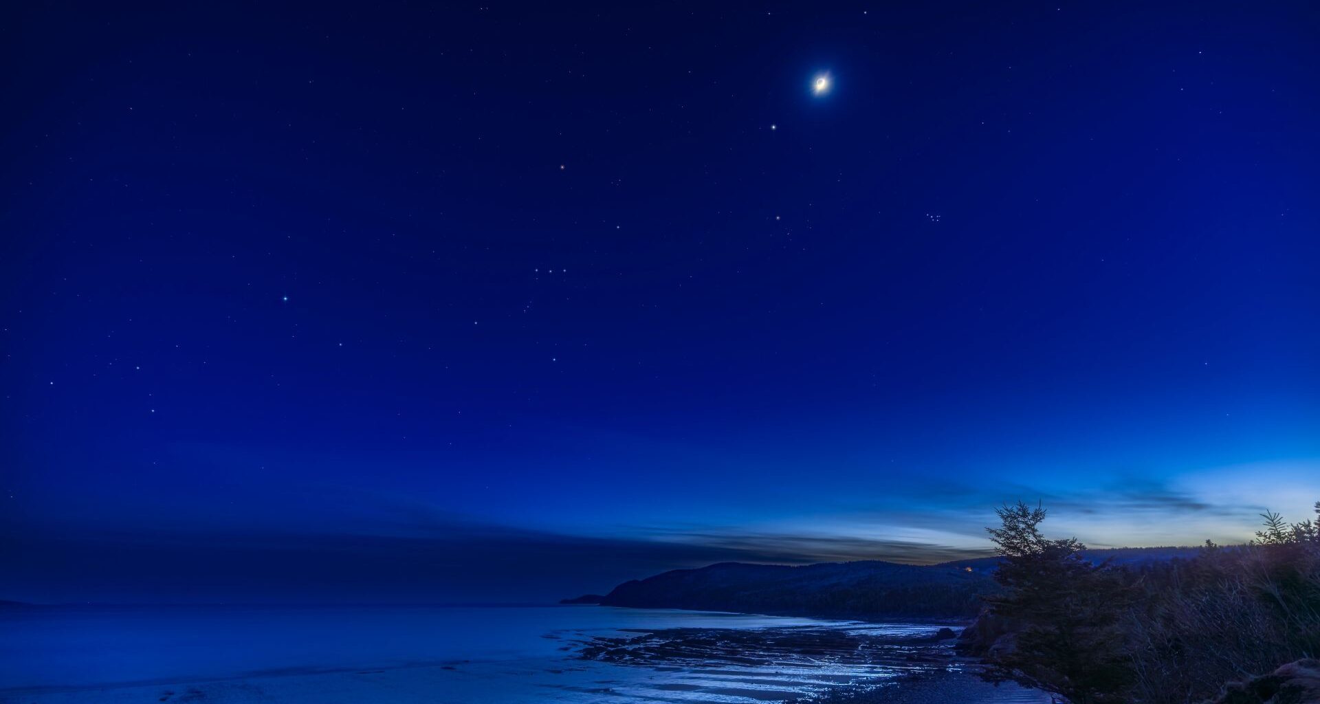 The moon is pictured glowing above the star-like form of Jupiter in a dark blue night sky above a snowy field bordering a road.