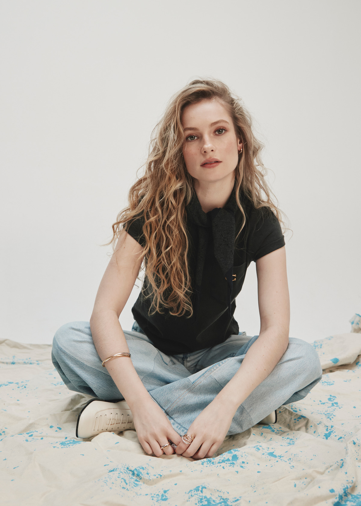 A woman with long wavy blonde hair wearing a black top and light jeans sits cross-legged on a canvas sheet with blue paint splatters, set against a plain light background in a studio environment.