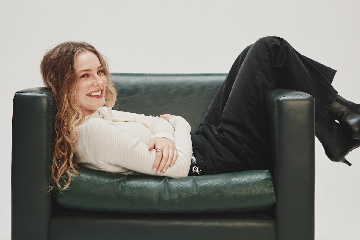 A smiling woman with long hair, wearing a light sweater and black pants, lounges sideways on a dark green armchair in a minimalist, well-lit studio environment.