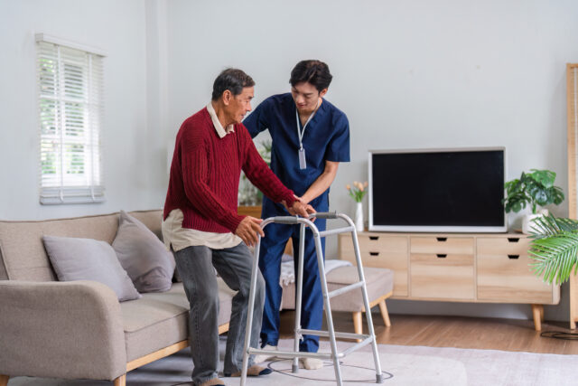 Caring nurse smiling while assisting elderly man with walker in sunny living room