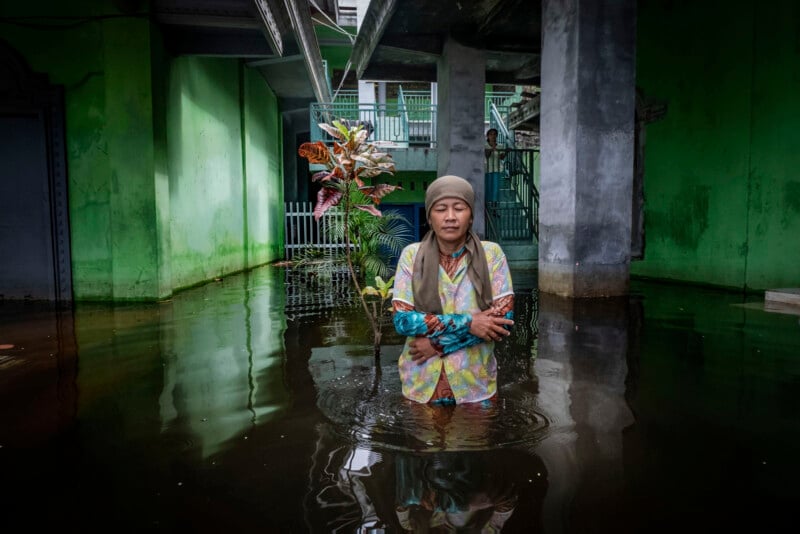 A woman wearing a headscarf and colorful clothes stands in knee-deep floodwater inside a building with green walls and potted plants, looking directly at the camera with arms crossed.