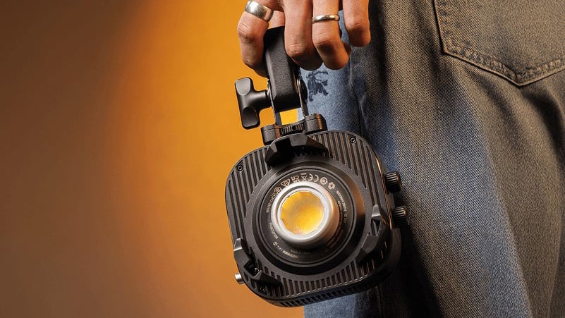 A person wearing jeans and rings holds a compact, portable LED studio light by its handle against an orange background.