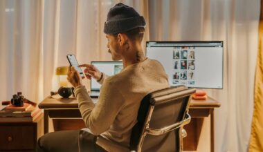A person wearing a beanie sits at a desk, using a smartphone. Two monitors, a laptop, camera, and headphones are on the desk. The room is warmly lit with curtains in the background.