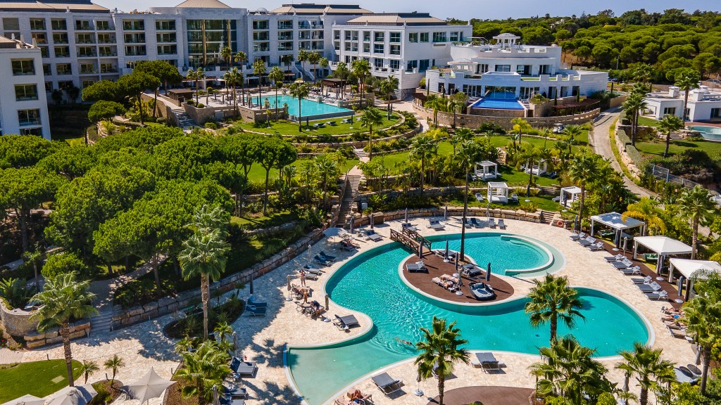 Resort pool with central deck, lounge chairs, cabanas, palm trees, and modern hotel building in a bright, tropical setting.