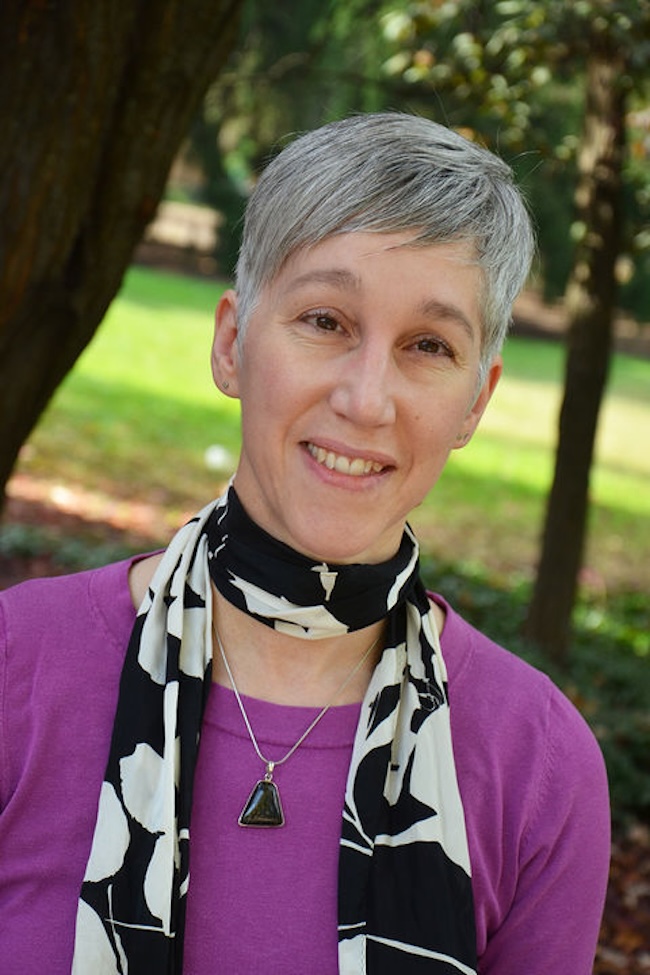Woman with short silver hair wearing a long black and white scarf and pendant. Some trees are behind her.