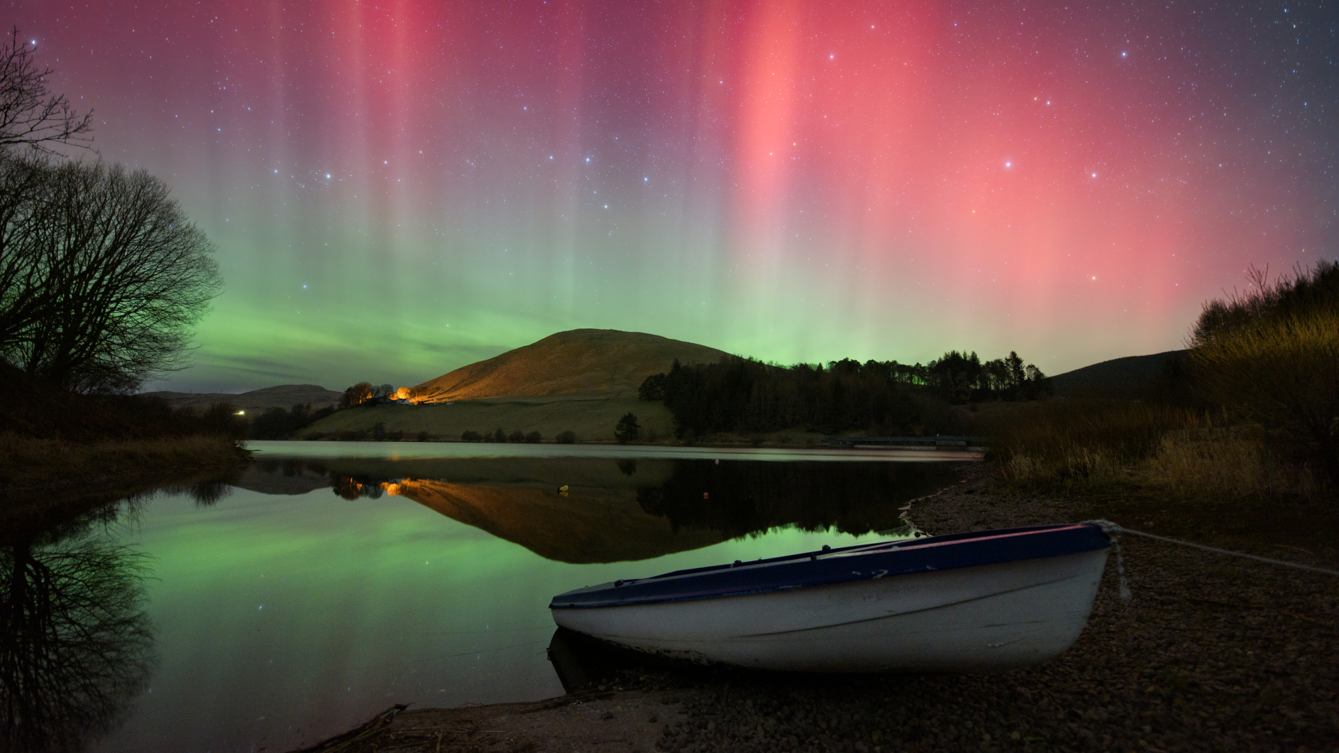 A small rowing boat is pulled onto the shore on the edge of a placid lake at night, as red and green auroras visible in the sky above reflect upon its surface.