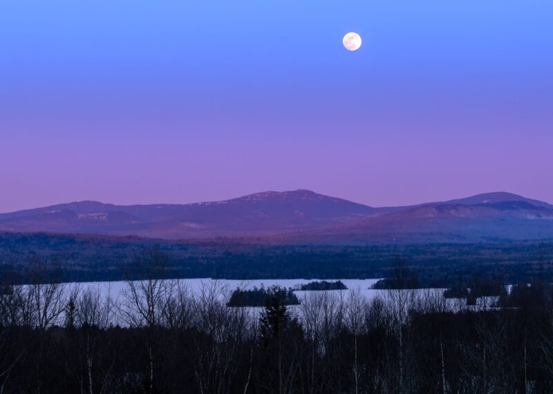 Full, white moon in a purple sky. There are some hills, a lake and trees below.