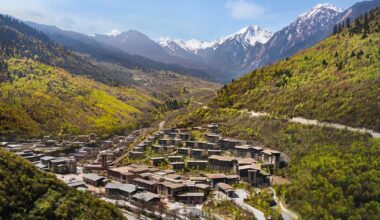 Mountain resort in lush valley with tiered buildings, forested hills, winding road, and snow-capped peaks under a clear blue sky.