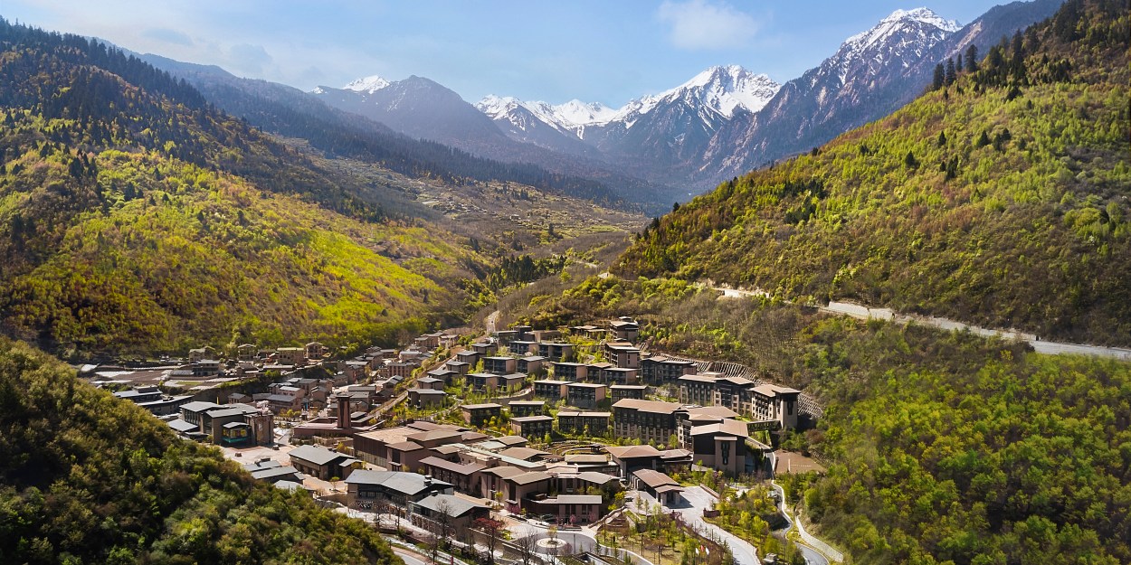 Mountain resort in lush valley with tiered buildings, forested hills, winding road, and snow-capped peaks under a clear blue sky.