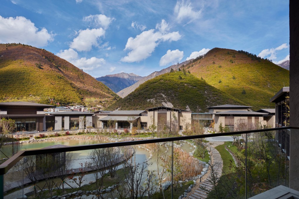 Modern buildings with glass windows, landscaped grounds, pond with walkway, and scenic hills under a blue sky.