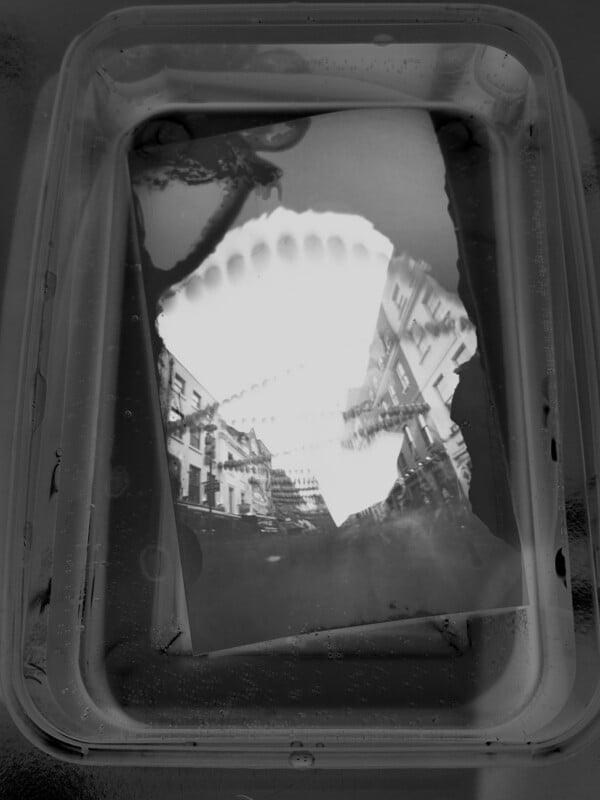 A black-and-white photo print is developing in a tray of liquid, showing a city street scene with buildings and trees reflected in the water. Edges of the photo are uneven and partially submerged.