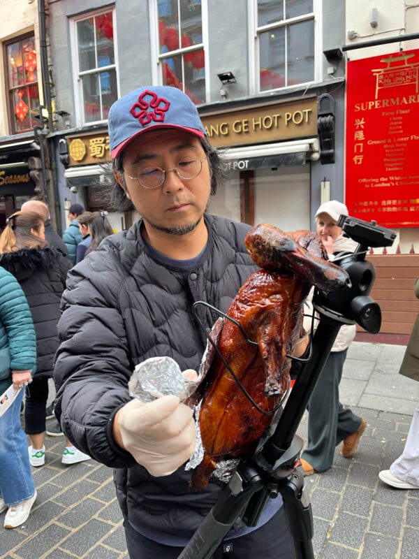 A man wearing glasses and a hat prepares a roasted duck on a metal stand outdoors, with people passing by and storefronts in the background.
