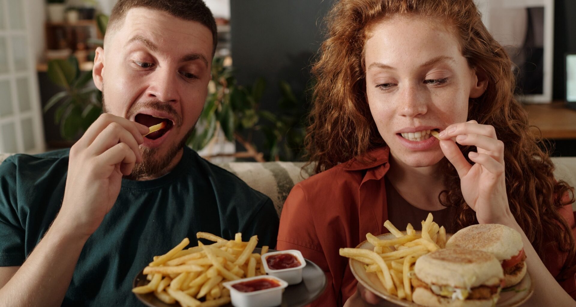 Young modern heterosexual couple eating french fries while sitting next to one another on couch in front of camera
