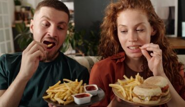 Young modern heterosexual couple eating french fries while sitting next to one another on couch in front of camera
