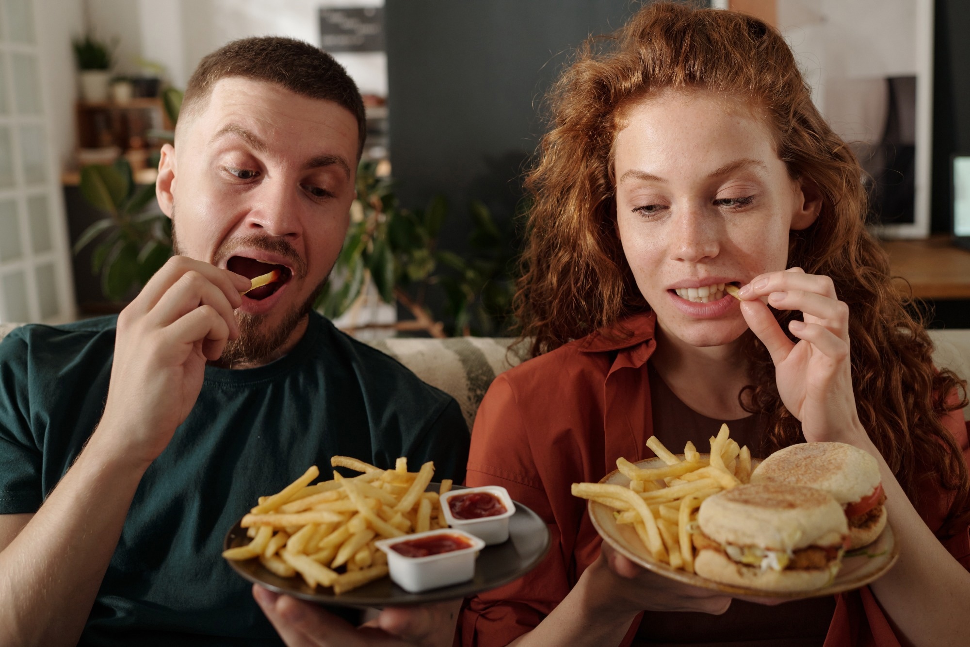 Young modern heterosexual couple eating french fries while sitting next to one another on couch in front of camera