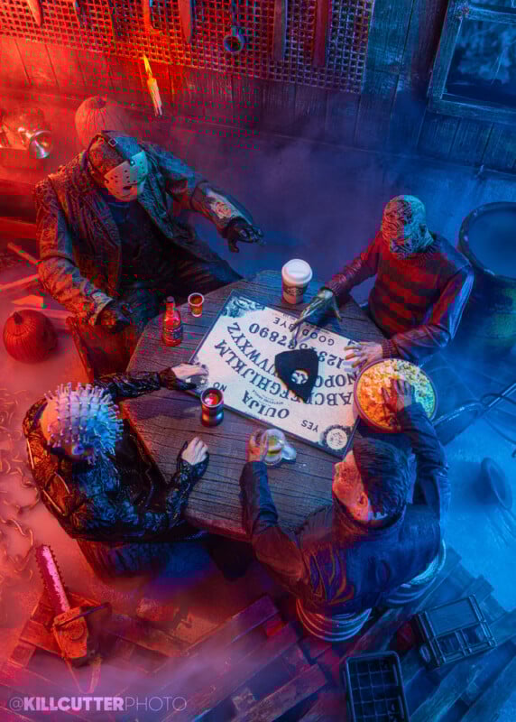 Four horror movie villains, including Jason Voorhees, Freddy Krueger, Pinhead, and Michael Myers, sit around a table with a Ouija board, snacks, and drinks in a spooky, dimly lit room decorated with weapons.