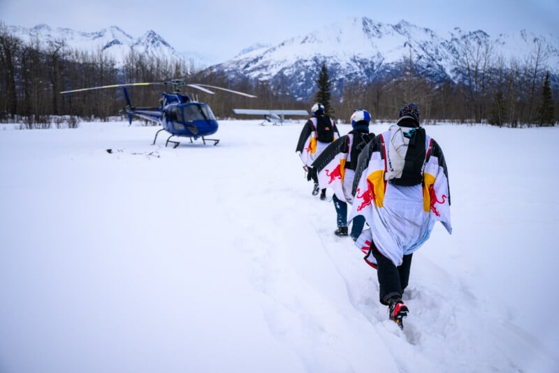 Three people in wingsuits walk through snow toward a blue helicopter, with snowy mountains and leafless trees in the background.