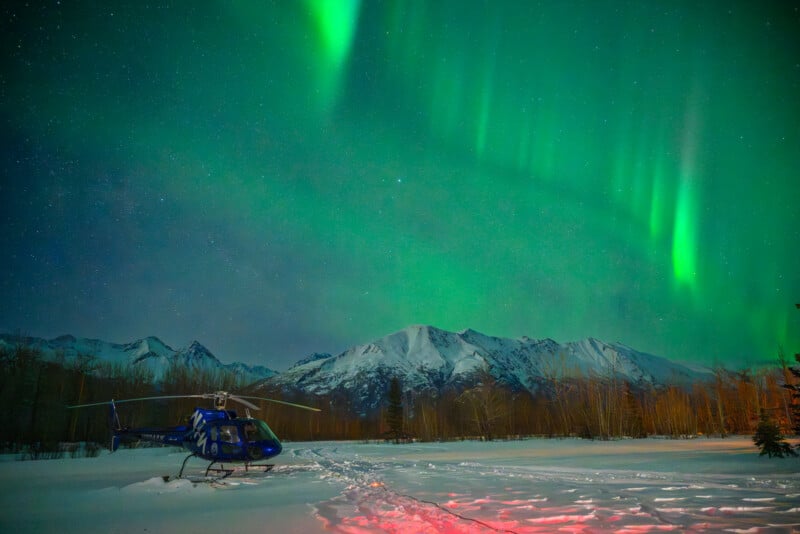 A blue helicopter sits on snowy ground beneath bright green aurora borealis lights in the night sky, with snow-covered mountains and leafless trees in the background.