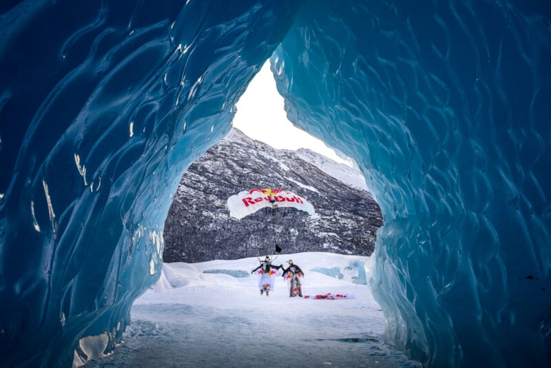 A view from inside a blue ice cave frames a skydiver landing on snowy ground, holding a parachute with the Red Bull logo. Snow-covered mountains are visible in the background.