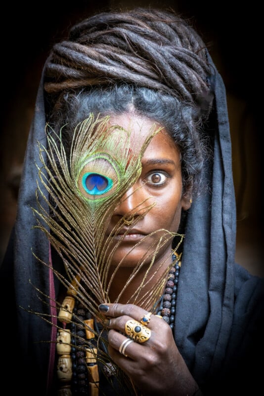 A person with long dreadlocks and layered jewelry holds a peacock feather partially covering their face, revealing one intense eye through the feather. The background is dark, highlighting their expressive features.
