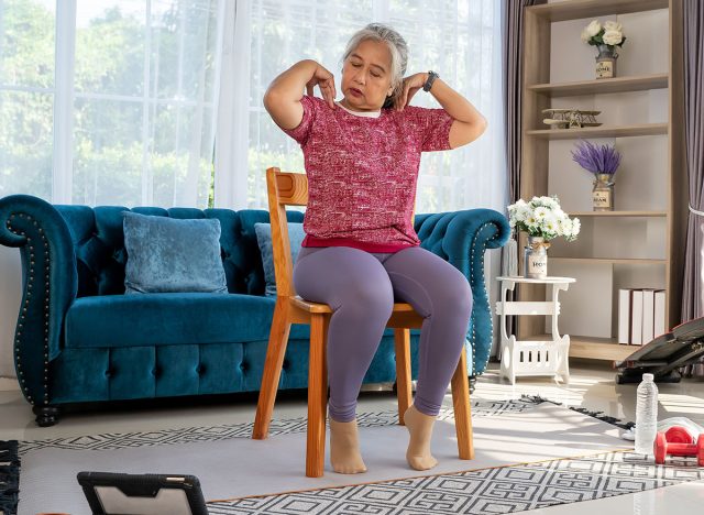 Senior woman sitting on a wooden chair and slowly rotate both shoulders, doing with training exercise online on tablet, workout online from home concept