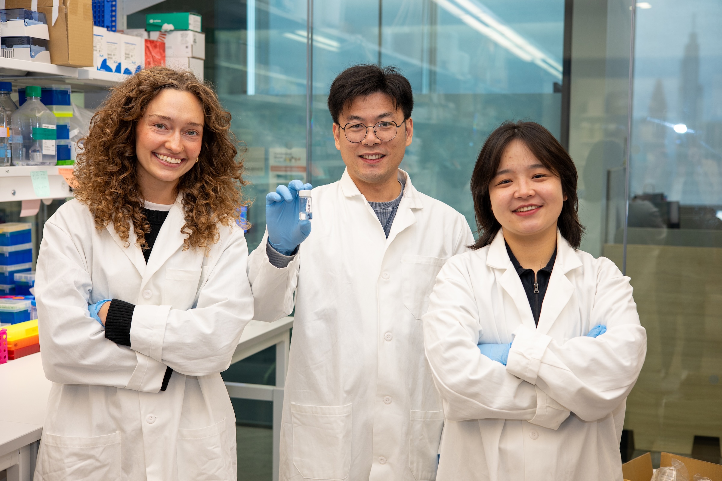 Three researchers in white lab coats stand side by side. The one in the middle holds up a small clear vial.